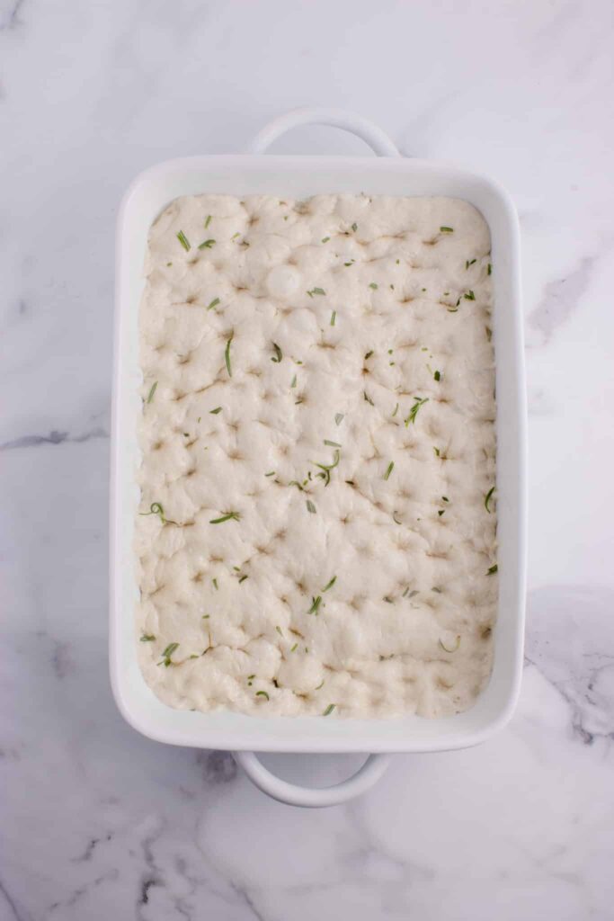 sourdough focaccia in white baking tray with dimples, rosemary, and flaky salt sprinkled on top ready to be baked