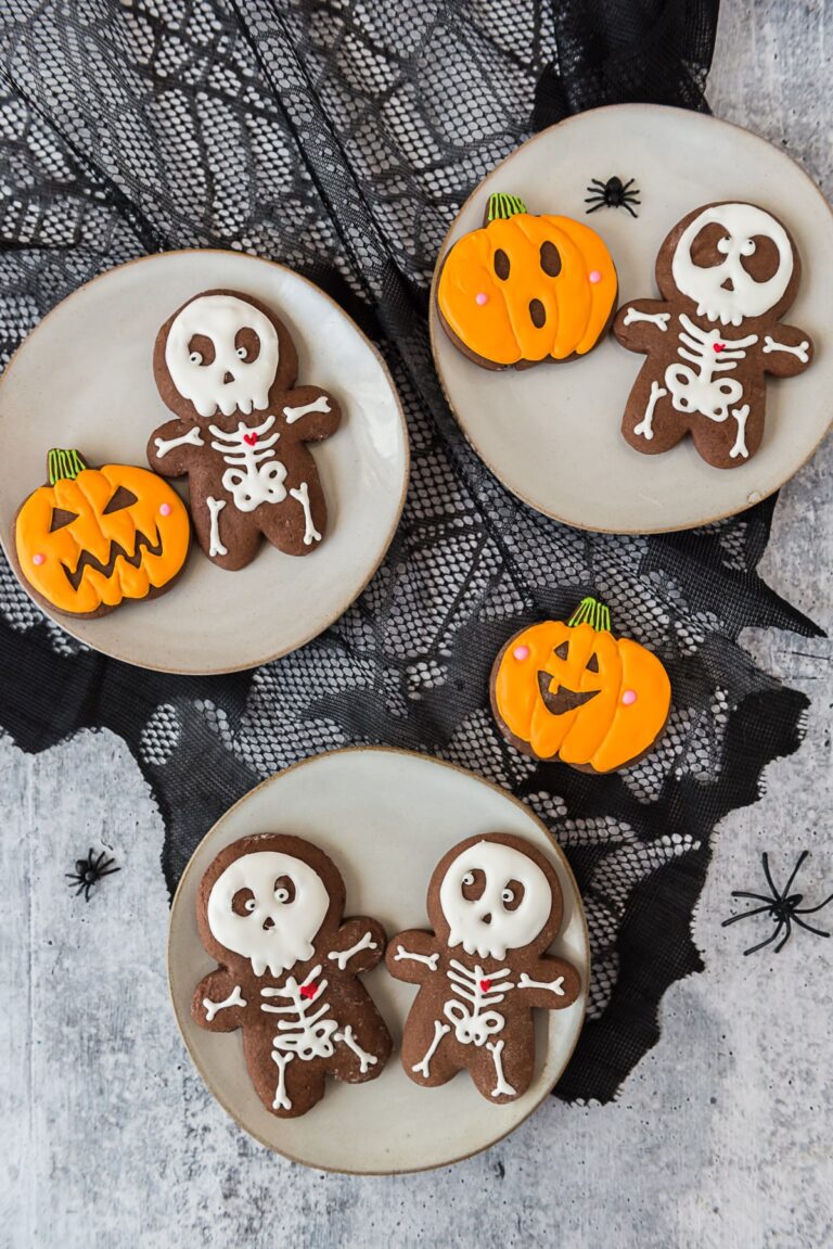 halloween decorated skeleton and pumpkin chocolate cut out cookies on three grey plates with plastic spiders scattered around
