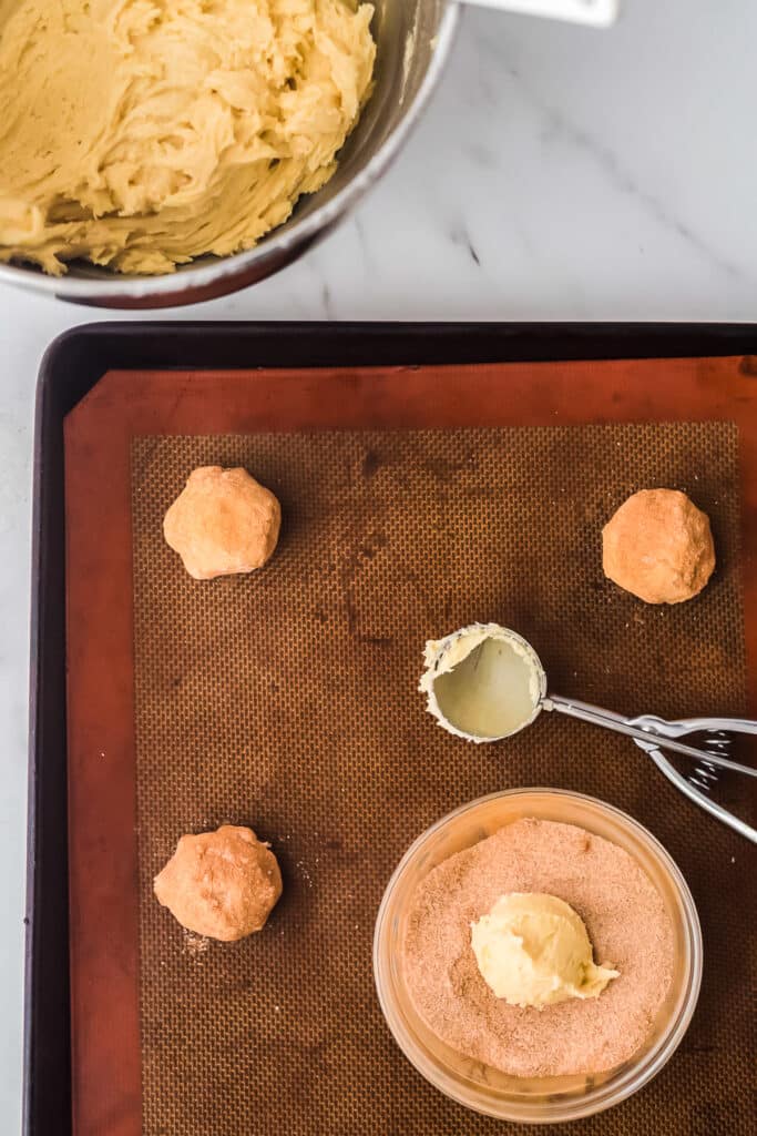 cookie dough being scooped into bowl of cinnamon sugar on cookie sheet