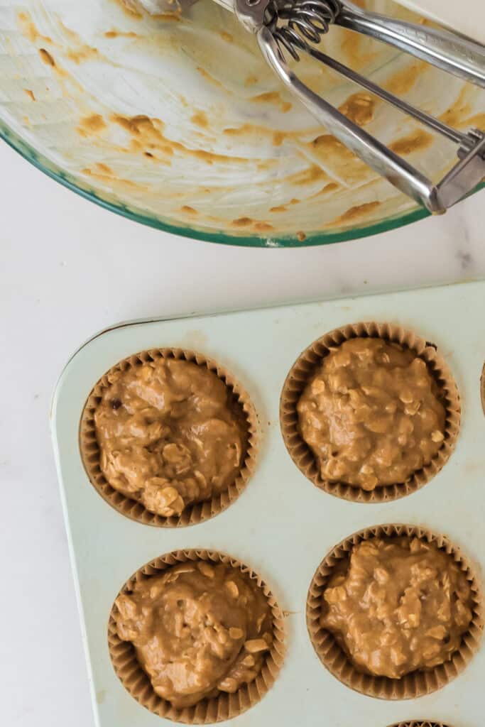 closeup of corner of muffin pan with four muffin wells filled with liners and batter and corner of empty mixing bowl