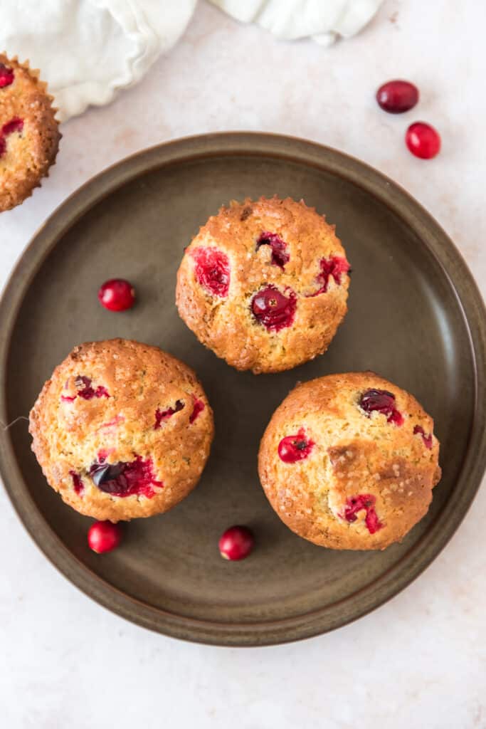 overhead view of three cranberry orange muffins on brown plate with cranberries scattered around