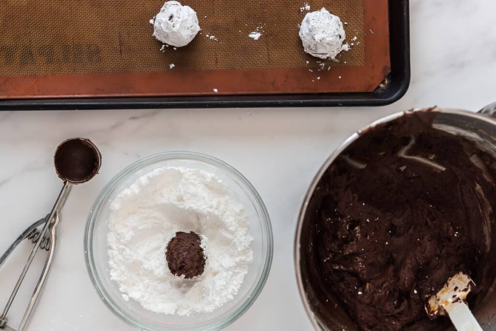scooping sourdough chocolate crinkle cookie dough into powdered sugar and baking sheet