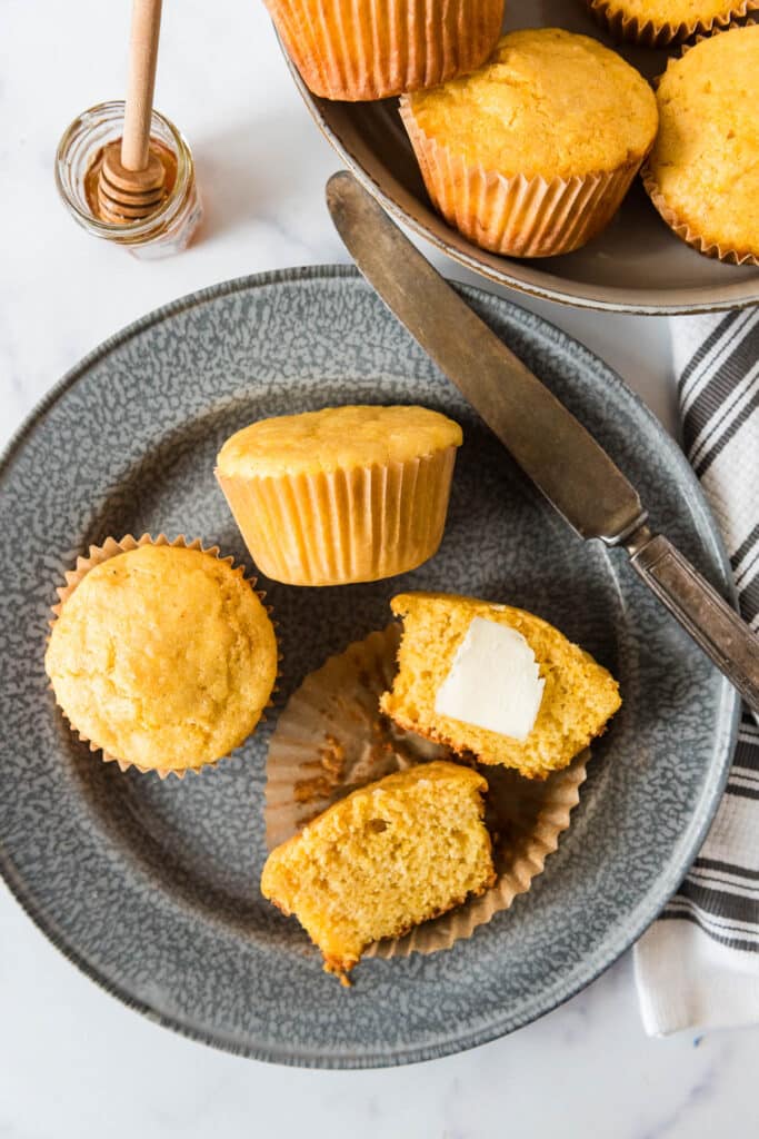 overhead view of three sourdough cornbread muffins on grey plate, one muffin is cut open and laying on its side with butter