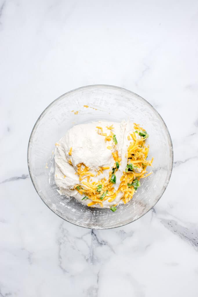 glass mixing bowl with bread dough and shredded cheese and jalapeno being mixed into dough