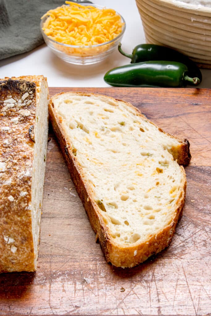 sourdough jalapeño cheddar bread with a slice on a wooden cutting board with cheese and jalapeno in background