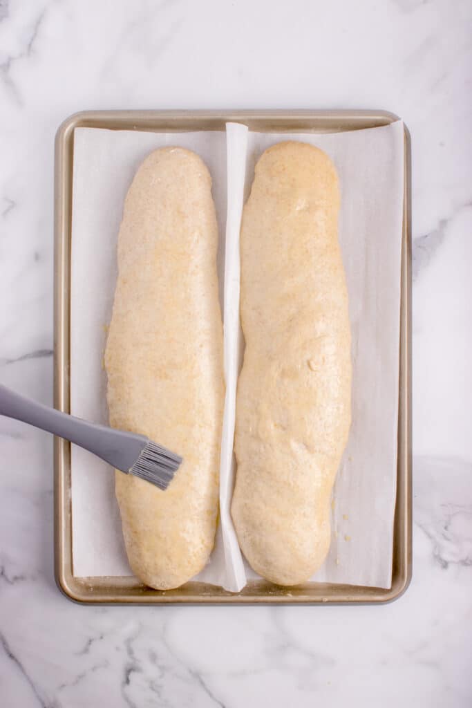 egg wash being brushed onto two loaves of French bread dough on baking tray