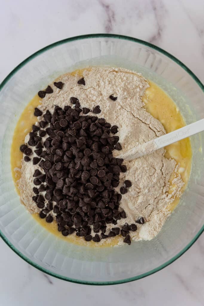 glass mixing bowl with  mixed wet ingredients, topped with flour, and chocolate chips ready to be stirred