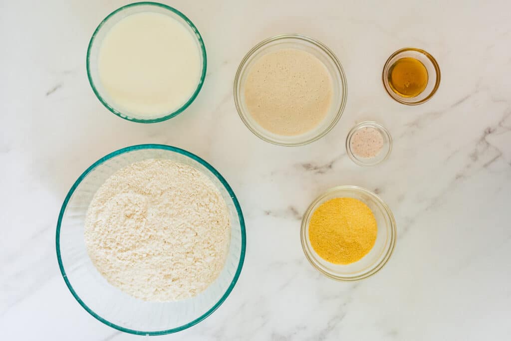 overhead of ingredients for sourdough English muffin bread in glass bowls on marble counter