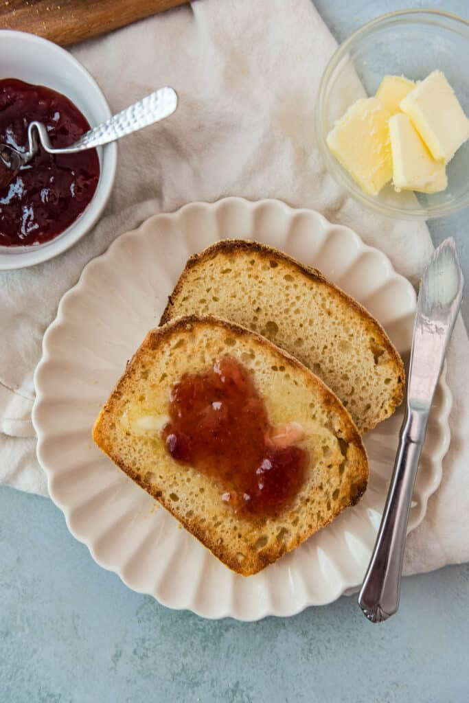 two slices of sourdough English muffin bread toasted with butter and jam on white plate with containers of butter and jam on the side