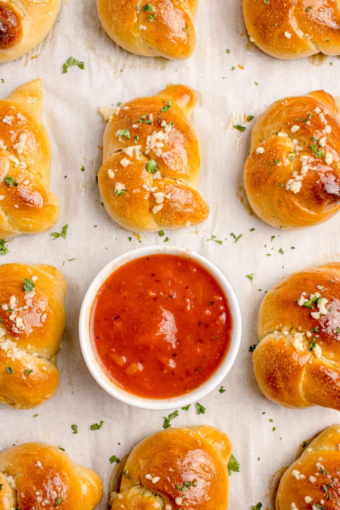sourdough garlic knots in a row on baking tray with one roll removed and replaced with bowl of marinara sauce