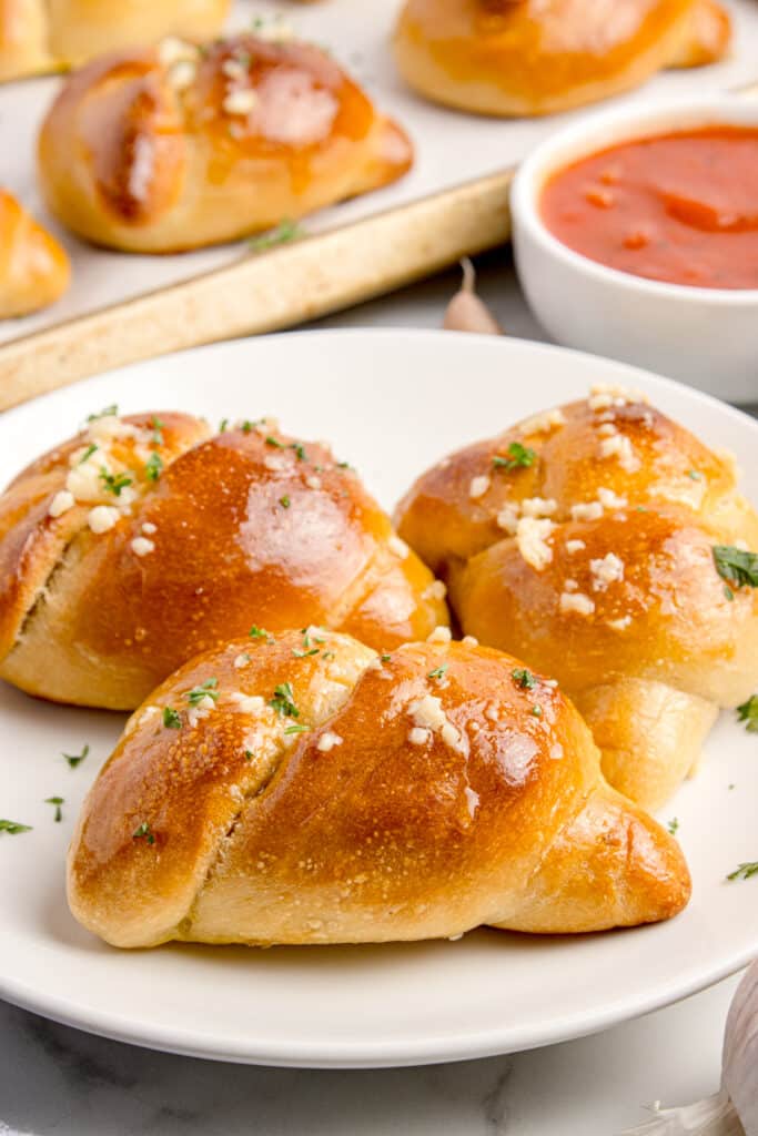 three sourdough garlic knots on white plate with bowl of marinara and tray with more rolls in background