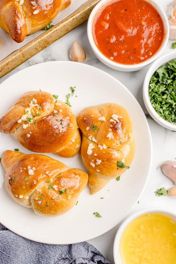 three sourdough garlic knots on white plate with bowls of marinara, parsley, and melted butter on the edges