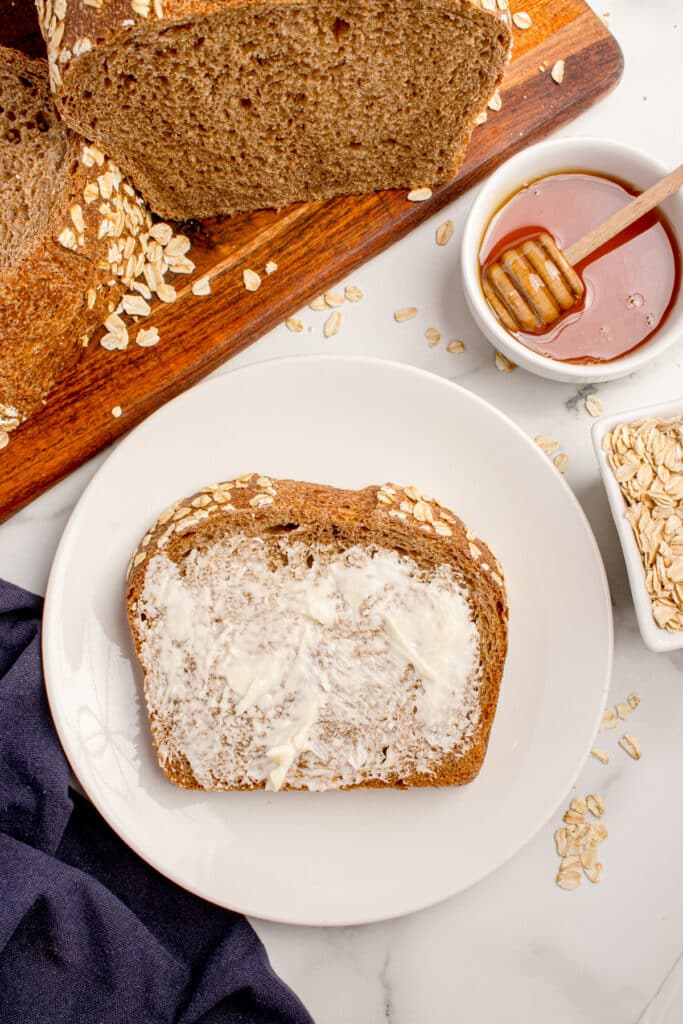 slice of sourdough brown bread on plate smeared with butter, with rest of loaf and jar of honey on the side
