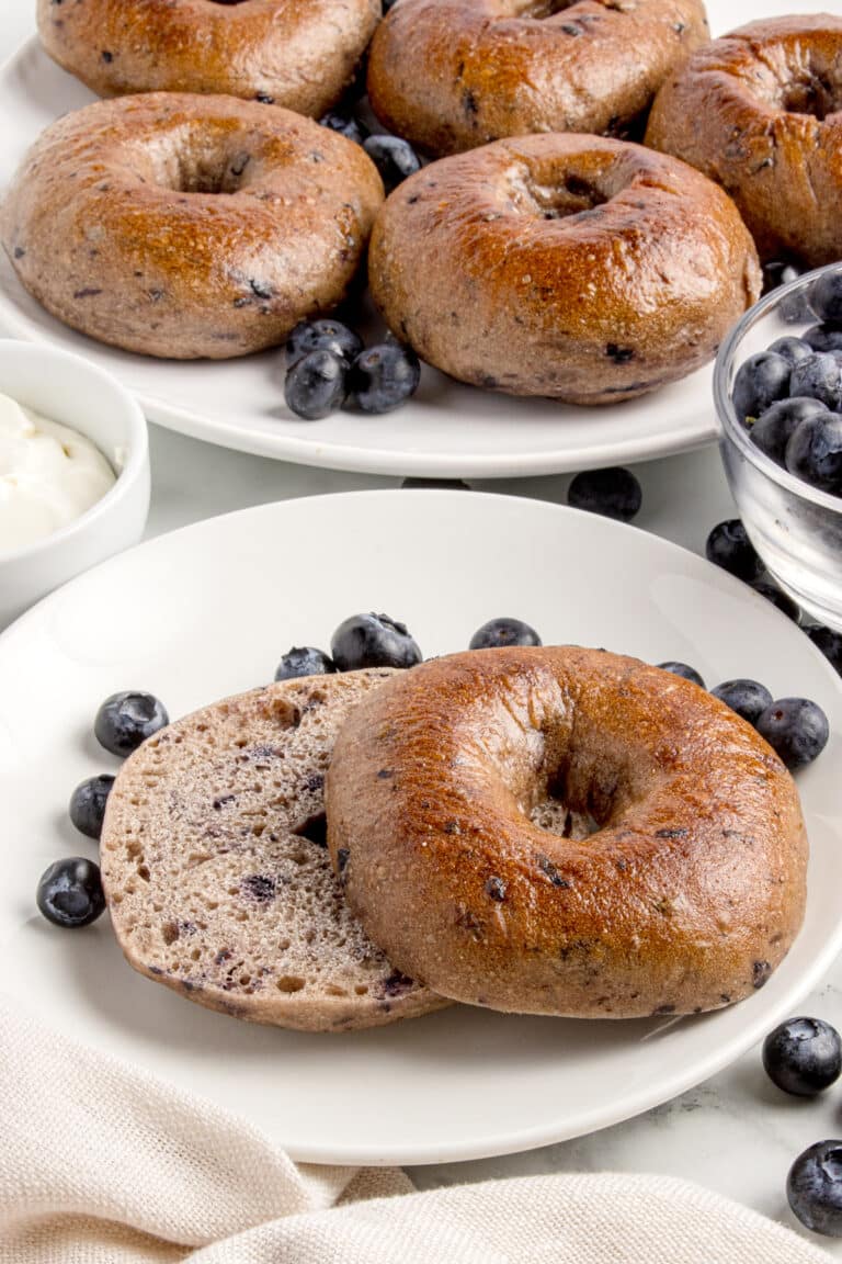 sourdough blueberry bagel cut in half on white plate with more bagels and blueberries surrounding it