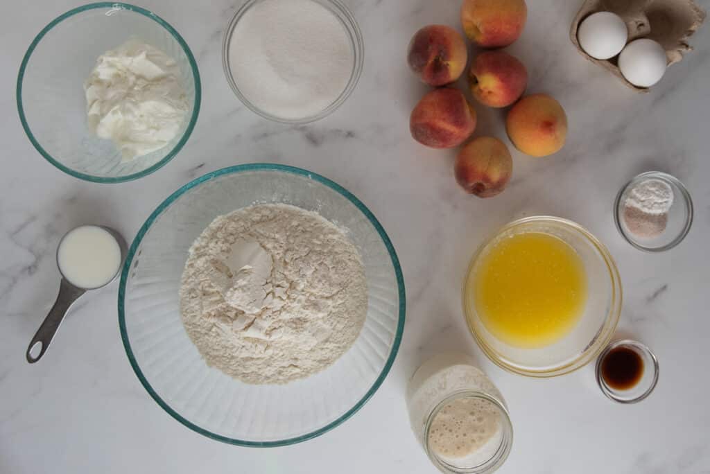 overhead view of ingredients for sourdough peach muffins in glass bowls on marble counter