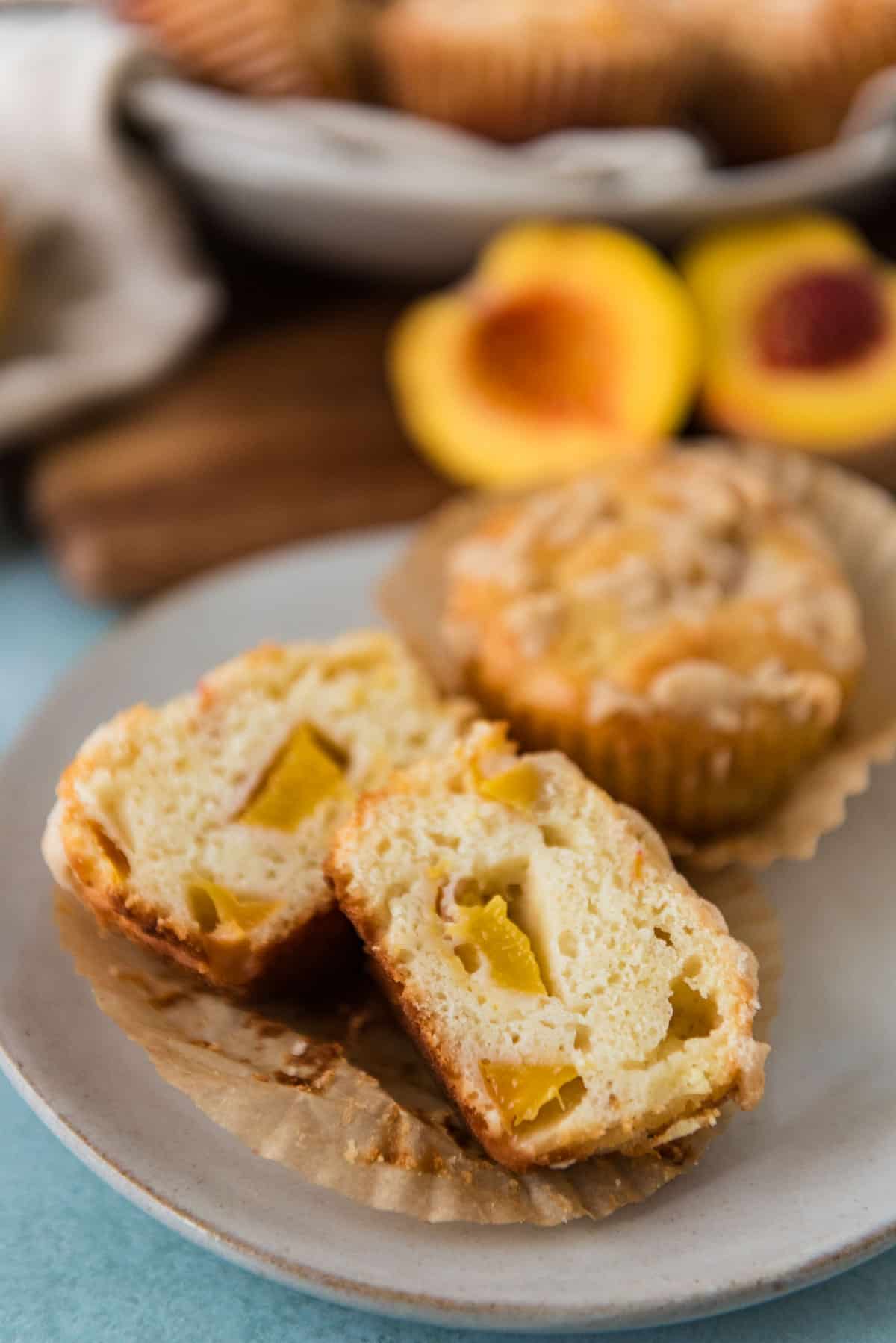 sourdough peach muffins on plate with one sliced to show inside texture with bowl of more muffins in background