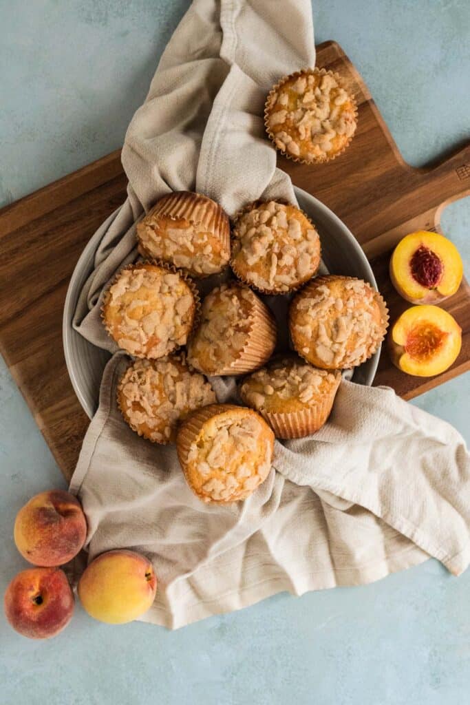 overhead view of bowl filled with peach sourdough muffins and napkin with fresh peaches surrounding it