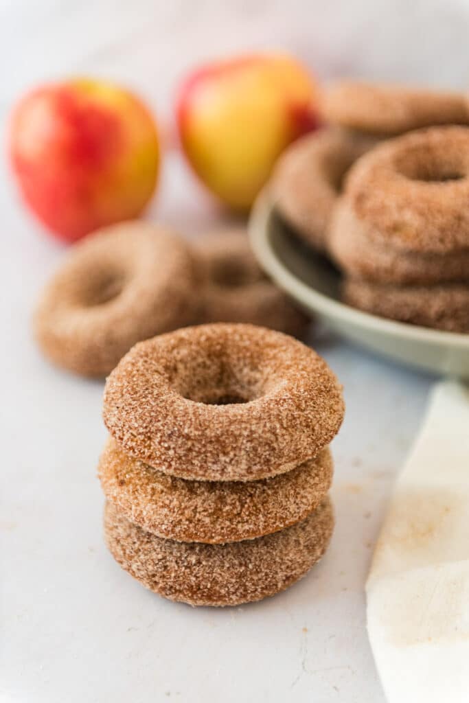 stack of three baked apple cider discard donuts with bowl of more donuts and apples in background