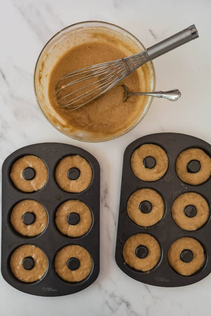 two donut pans filled with cider donut batter next to bowl of batter with whisk and spoon