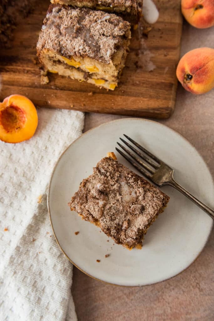 overhead view of peach sourdough coffee cake on grey plate with fork, beside cutting board with more cake and peaches