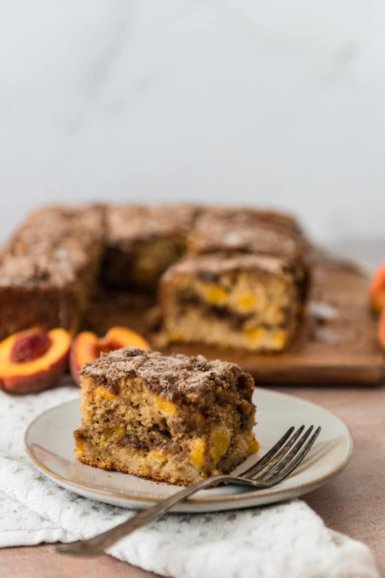 slice of peach sourdough coffee cake on grey plate with fork and rest of cake and sliced peaches in background