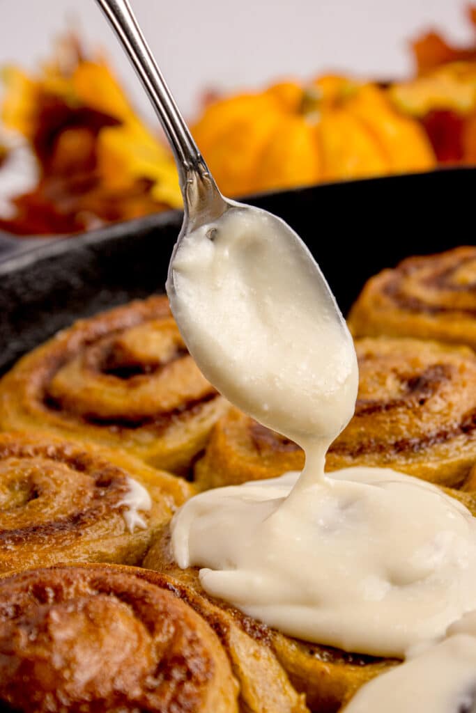 spoon drizzling cream cheese frosting on sourdough pumpkin cinnamon rolls
