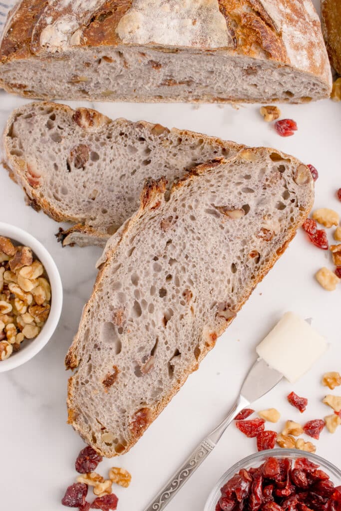 loaf of cranberry walnut sourdough bread with two slices stacked in front and scattered walnuts and dried cranberries