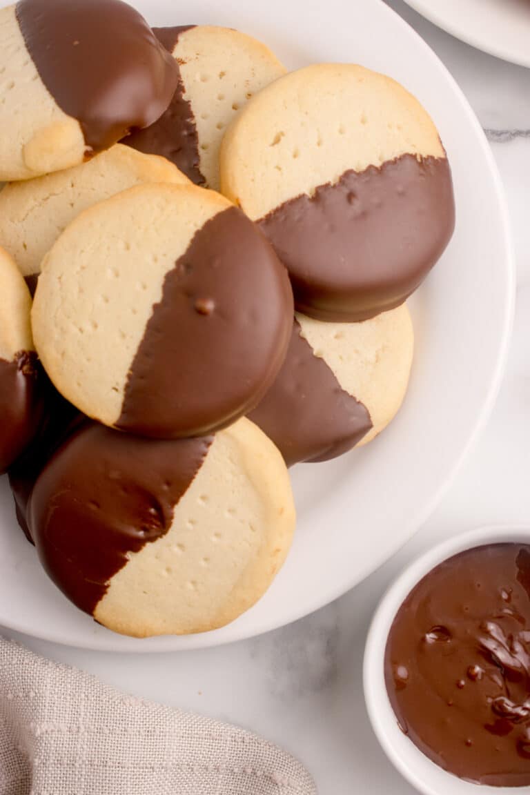 overhead view of white platter stacked with sourdough shortbread cookies dipped halfway with chocolate and small bowl of melted chocolate beside